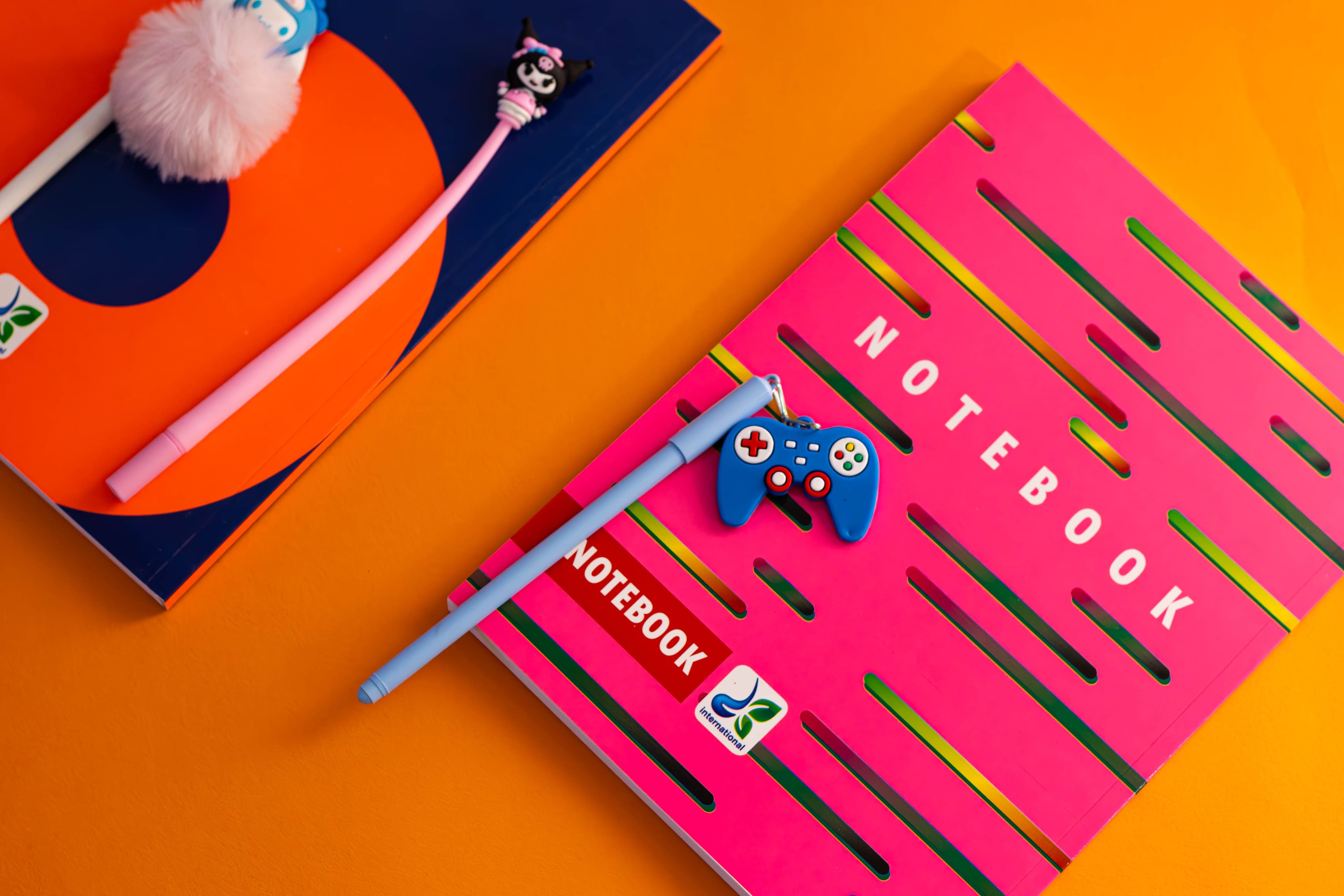 Two colorful notebooks on an orange background — a navy notebook with an orange circle design and a hot-pink notebook with rainbow stripe cutouts — accompanied by two fuzzy character-topped pens with a pink pom-pom and a blue controller-shaped keychain