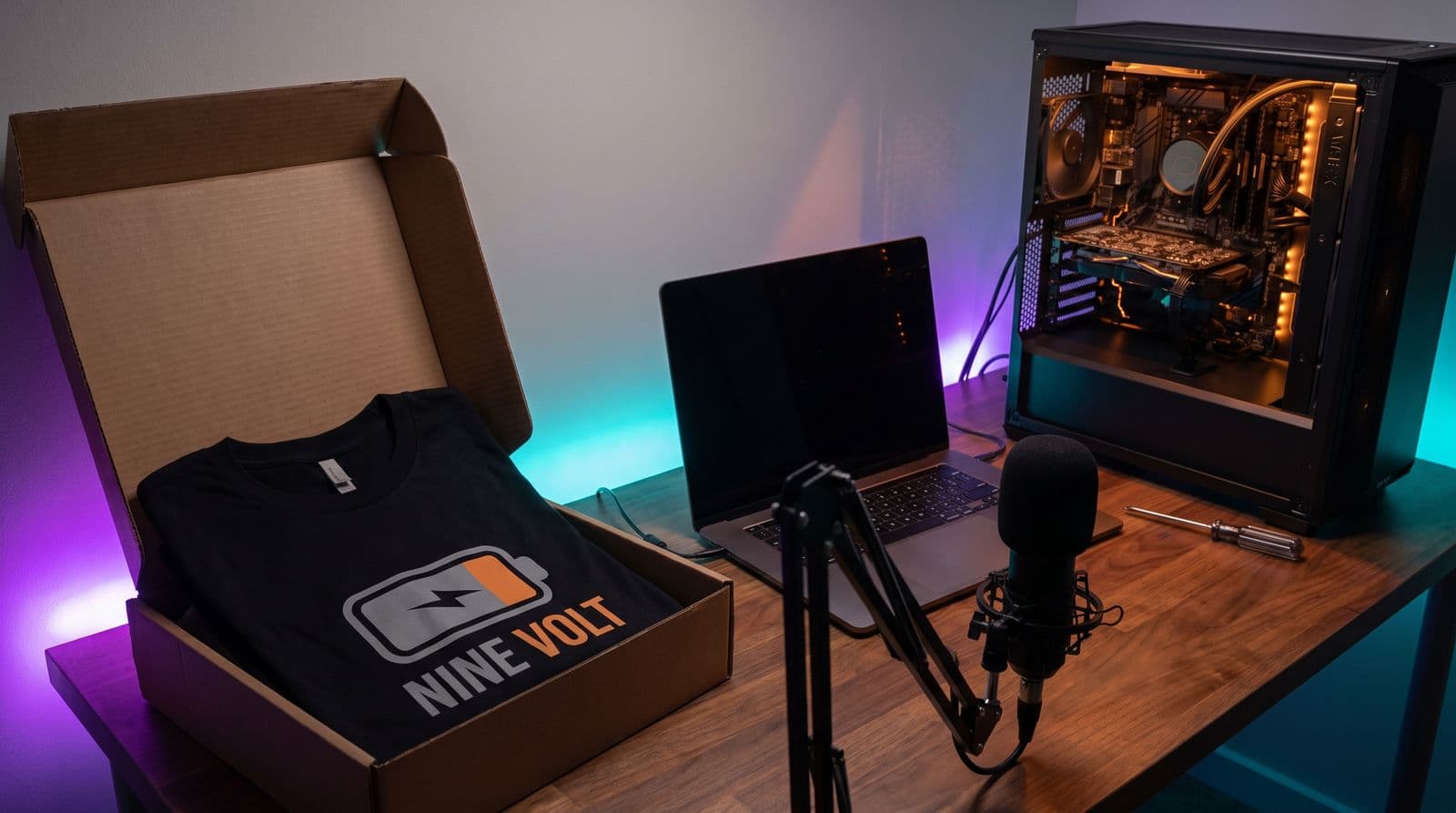 A streaming desk in purple and teal light: on the left a sealed mailer box with a folded black tee printed with the invented channel name Nine Volt, in the middle a laptop with a dark screen and a microphone, and on the right an open PC case with its components exposed and a screwdriver beside it.