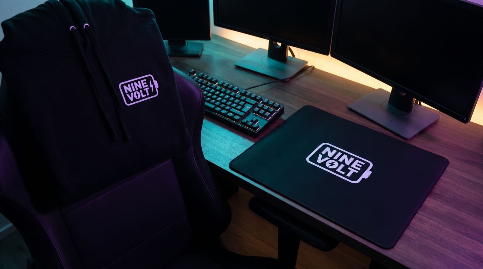 A black hoodie and a mousepad featuring the NINE VOLT brand logo sit on a gaming desk illuminated by purple and teal ambient lighting.