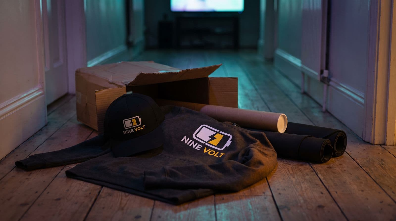 A courier parcel sits open on a hallway floor, revealing a branded hoodie and cap from the channel NINE VOLT.