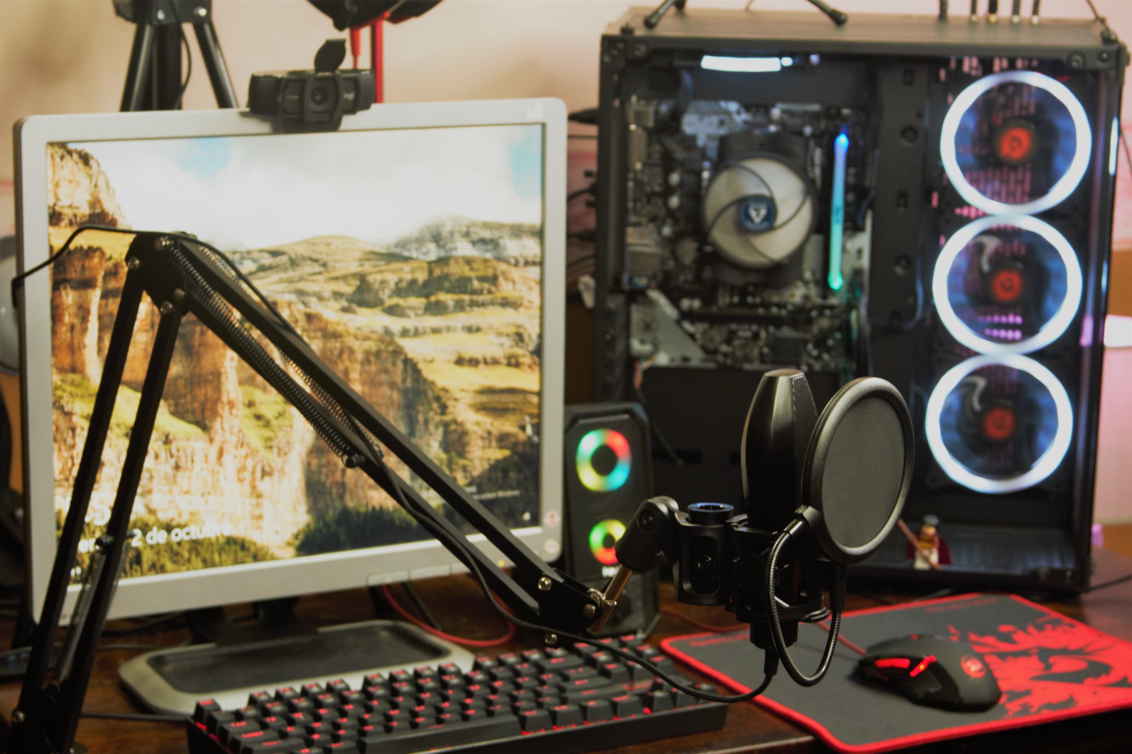 A gaming and streaming desk setup with an open PC case showing RGB fan lighting, a boom-arm microphone with pop filter, a backlit red mechanical keyboard, RGB speakers, and a monitor displaying a canyon landscape image