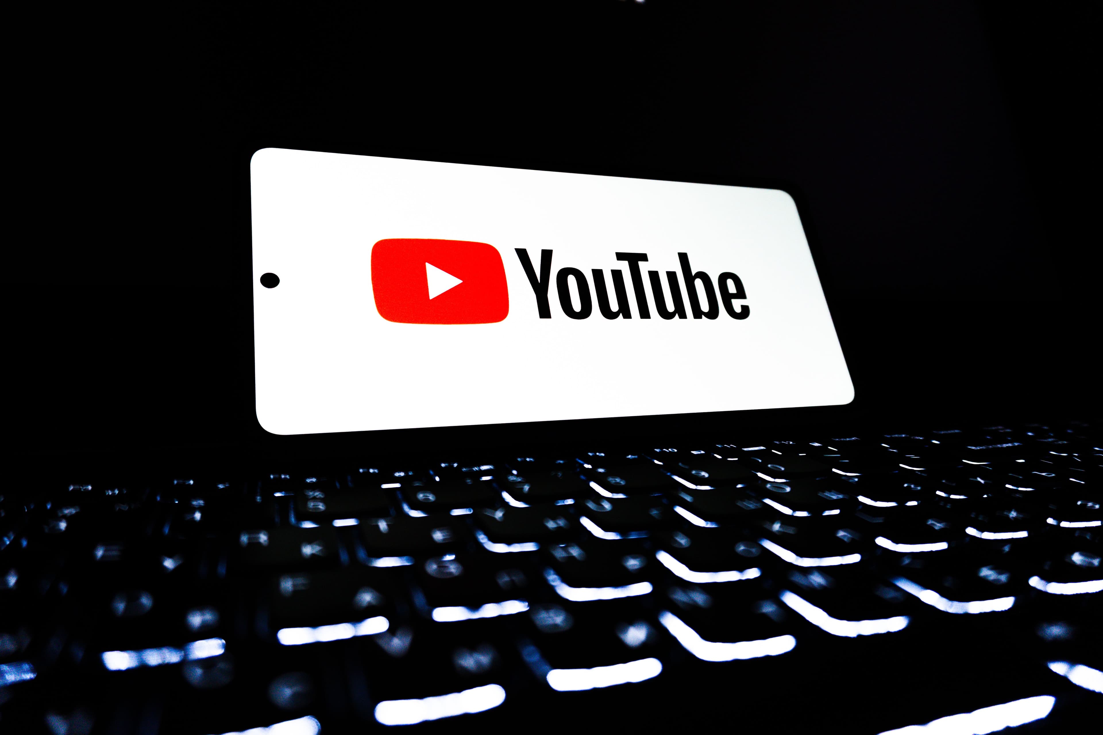 Smartphone screen displaying the YouTube logo standing upright behind a backlit computer keyboard, both against a black background