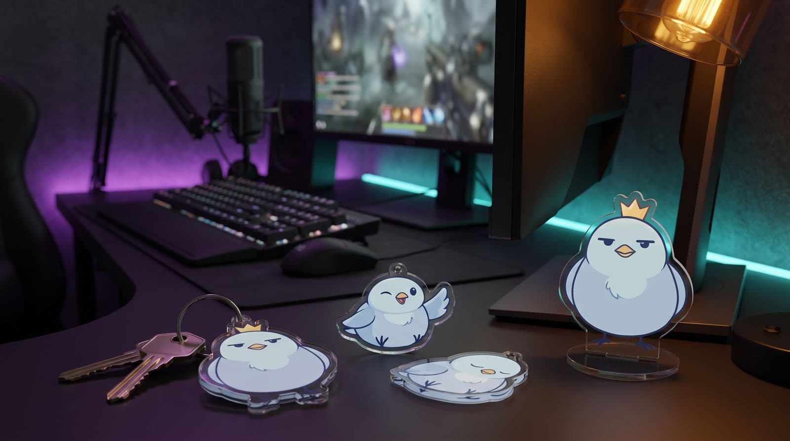 Clear acrylic merch of a round blue cartoon bird with a crown on a streaming desk: a keychain on a set of keys, two loose charms and a small standee, with a keyboard, microphone and glowing monitor behind.