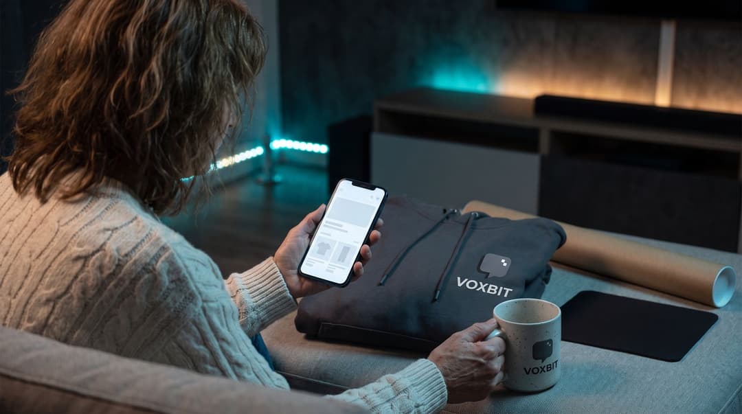 A creator in relaxed knitwear sits on a sofa in a dimly lit room, checking her VOXBIT-branded storefront on a smartphone with a matching hoodie and mug resting nearby.