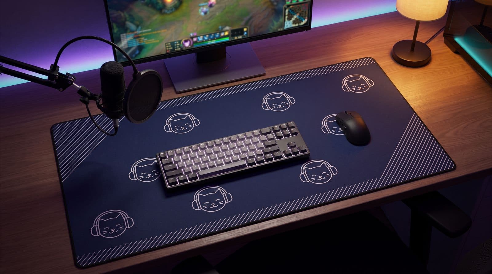 A navy desk mat printed with a repeating cat-in-headphones mark and thin diagonal lines along its edges, a keyboard and mouse on top, on a streaming desk with a microphone, a monitor and purple LED light.