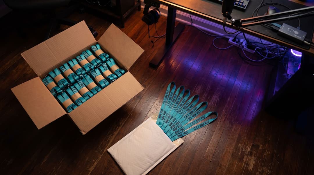 Overhead view of a streaming-room floor: a large open box packed with banded bundles of teal Nine Volt lanyards next to a padded mailer with ten of the same lanyards fanned out on top, the desk legs and cables beyond.