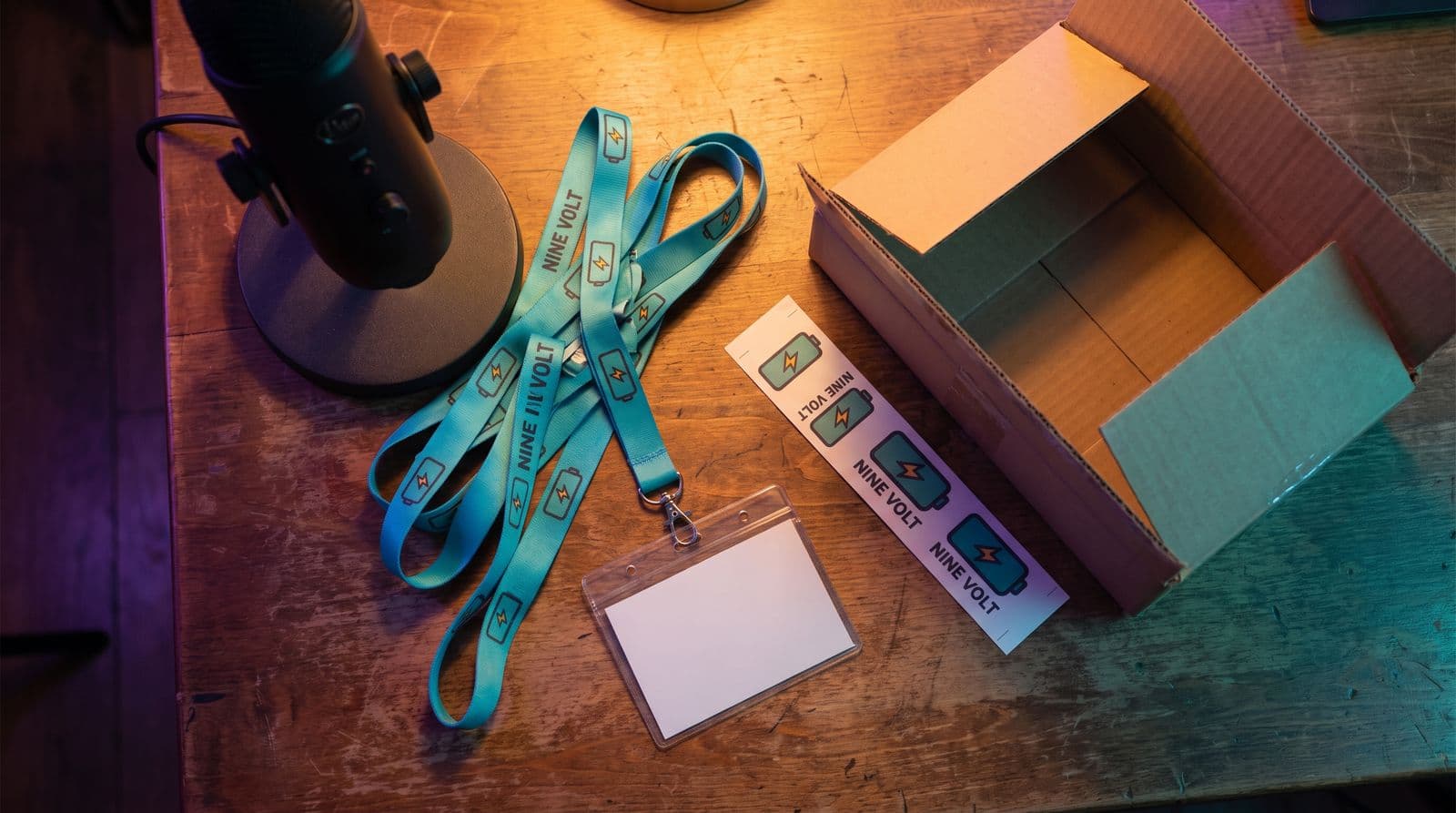 Overhead view of a streaming desk: a tangle of teal custom lanyards printed with the invented channel name Nine Volt, one clipped to a blank badge holder, a printed design proof strip, an open shipping box and the base of a microphone, in warm and purple light.