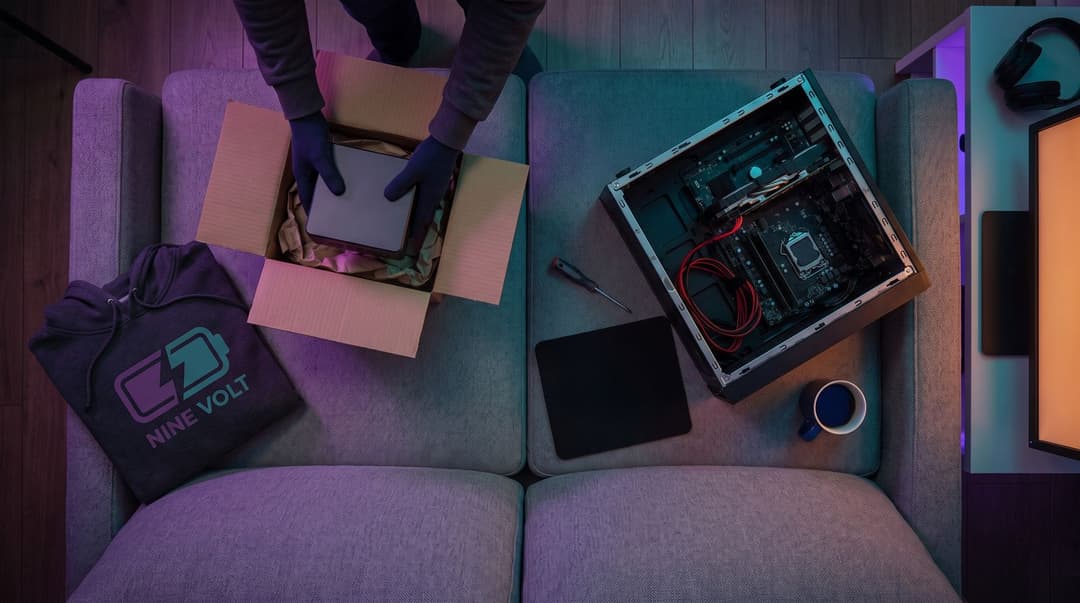 Overhead view of a sofa in evening light: hands lifting a small complete mini PC out of its box on one cushion, an open empty PC case with a motherboard, cables and a screwdriver on the other, and a folded Nine Volt hoodie and a mug beside them.