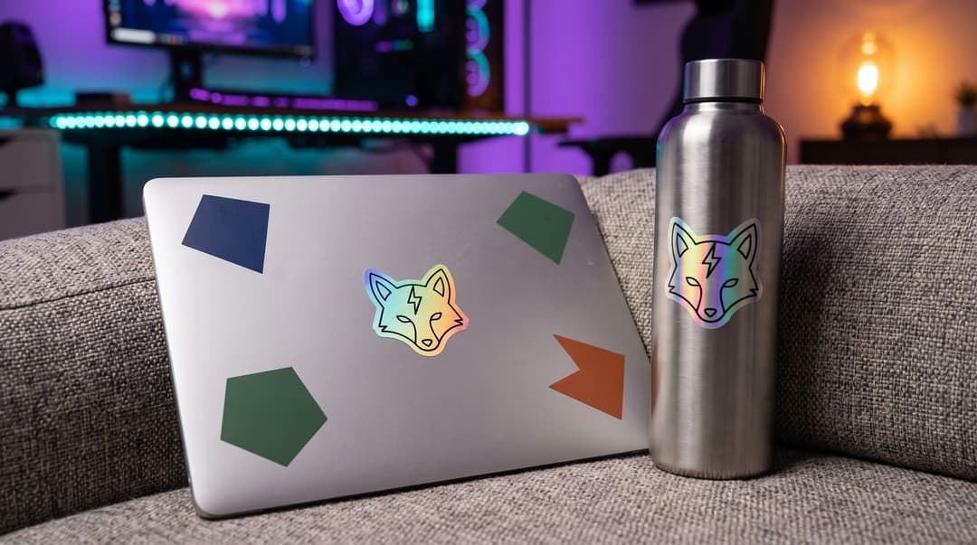 A closed laptop on a sofa with a holographic fox sticker among plain coloured shapes, and a steel water bottle with the same sticker, a streaming setup glowing purple and teal behind.