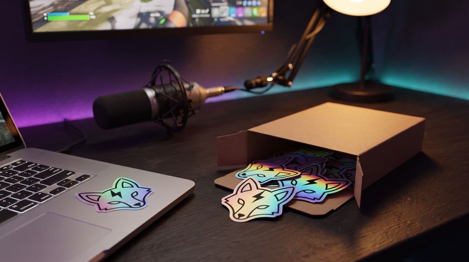 Holographic die-cut stickers of a fox head with a lightning bolt spilling from a small cardboard mailer on a dark streaming desk, one already stuck on the laptop beside a microphone and a glowing monitor.
