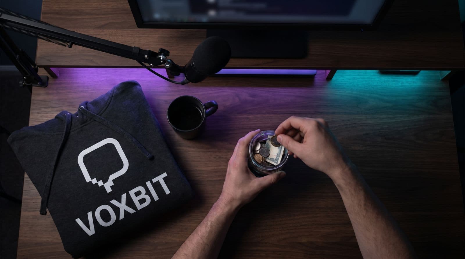 Hands dropping coins and a folded note into a small glass jar on a streaming desk lit purple and teal, a folded charcoal hoodie for an invented channel called Voxbit beside the mug and mic arm.