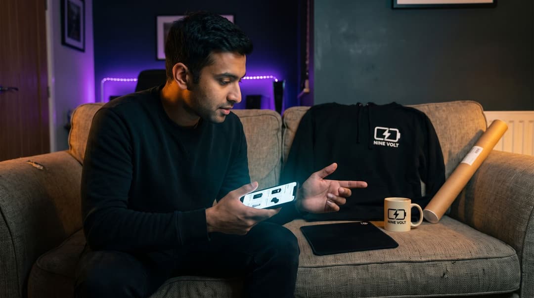 A man sits on a sofa in a dimly lit room, comparing his smartphone screen to a NINE VOLT branded hoodie and mug to see if his merch designs match his community's preferences.