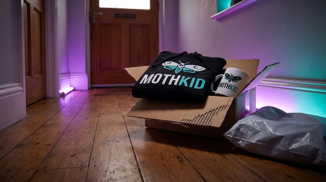 A black hoodie and a white mug featuring the MOTHKID moth logo sit inside an opened courier parcel on the floor of a dimly lit hallway.