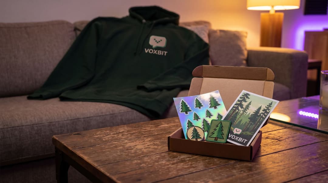 A small open mailer on a coffee table holding a holographic pine-tree sticker sheet, an embroidered pine patch, a round pin and a small forest print, a forest-green Voxbit hoodie draped over the sofa behind in lamp light.