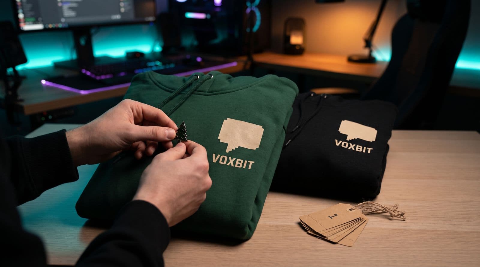 Hands pinning a small pine-tree enamel pin onto a forest-green members-edition hoodie for an invented channel called Voxbit, the public black hoodie folded beside it and a stack of numbered kraft tags on a streaming desk lit purple and teal.