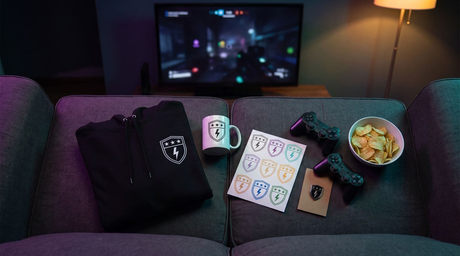 A folded black hoodie, a white mug, a sticker sheet and an enamel pin, all printed with the same shield-and-lightning emblem, laid out on a sofa with two controllers and a bowl of crisps in front of a TV.