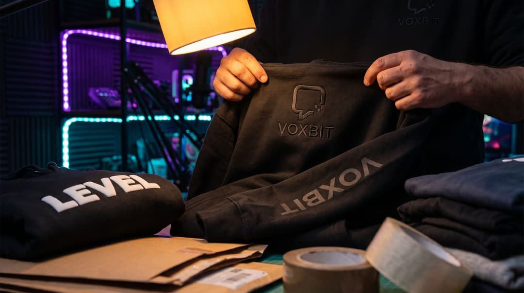 Hands holding a black hoodie under a desk lamp to show tone-on-tone black embroidery of the VOXBIT mark on the chest, the channel name printed down one sleeve, and a second hoodie beside it with the word LEVEL in raised white puff print, mailers and tape on the desk.