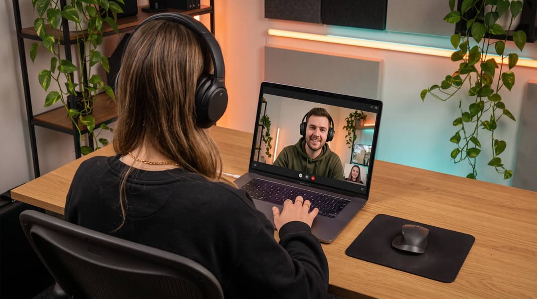 Creator in headphones on a video call with a collaborator, seen over her shoulder at a home desk